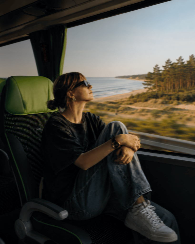 Nuotrauka su mergina sėdinčia autobuse ir žiūrinčia pro langą į jūrą
